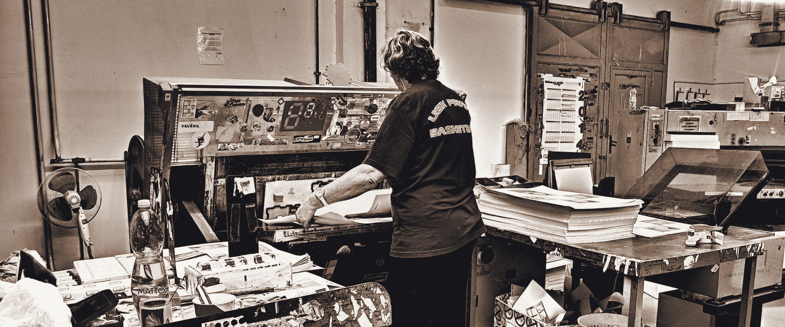 A print shop worker feeding printed sheets through a cutter, stacks of finished work on the bench beside her.